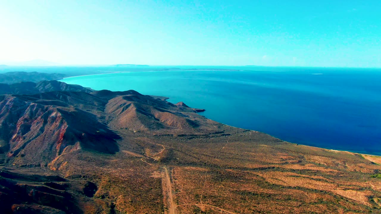 High aerial frame over Mulege, Baja California Sur, looking past brown desert mountains and pale road lines toward a bright blue coastal bay with no visible people.