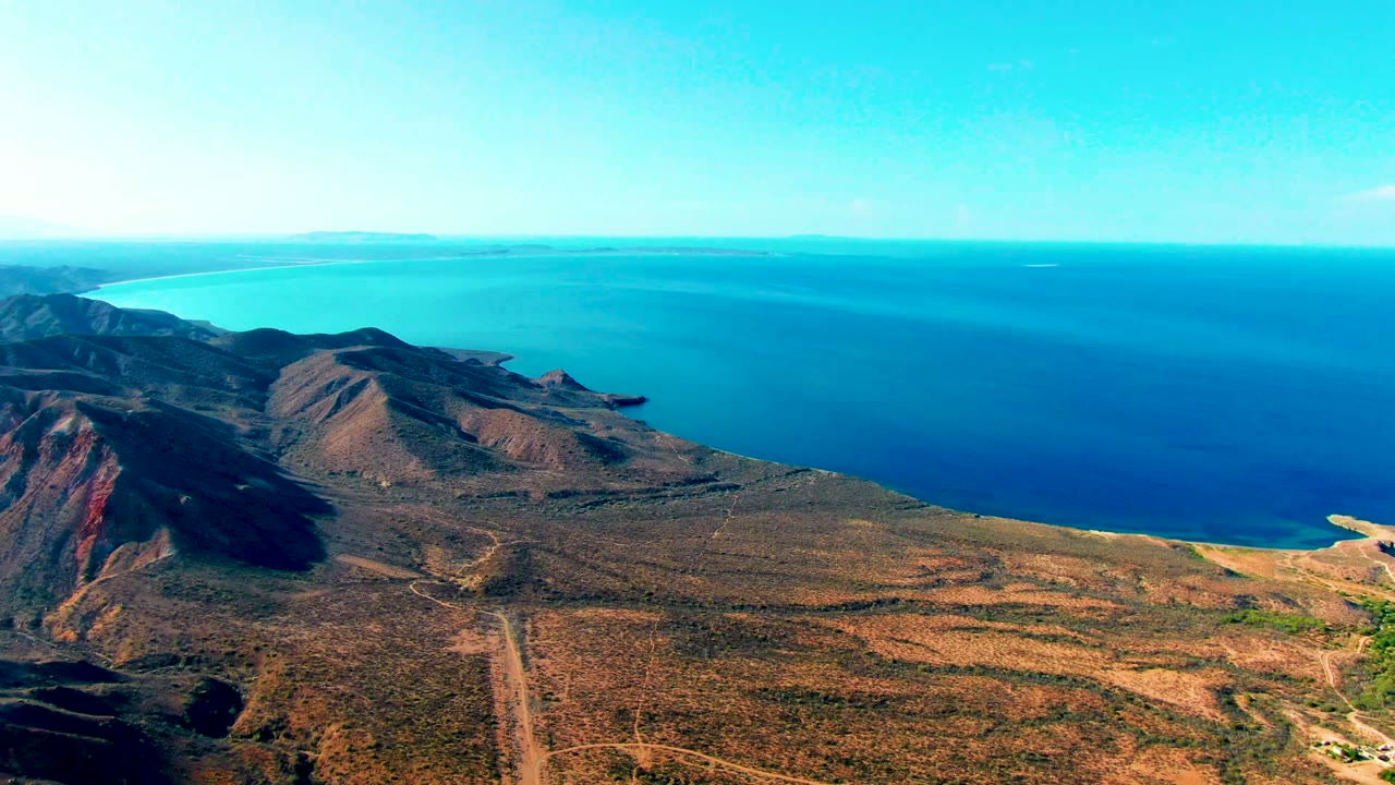 Aerial frame over Mulege, Baja California Sur, with dry mountains at left, scrubland and a dirt road below, blue ocean beyond, and no visible people.