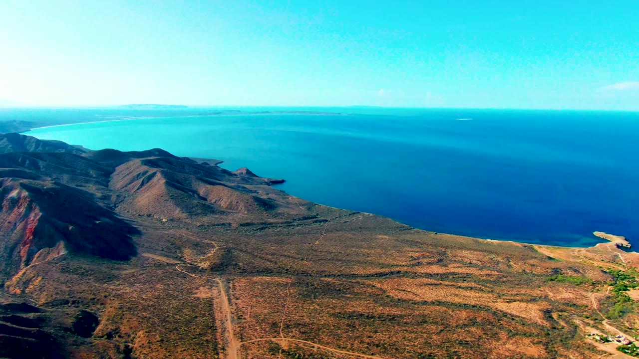 Mulege aerial frame with rocky brown hills at left, open scrub plain across the lower frame, bright blue sea in the distance, and no visible people.