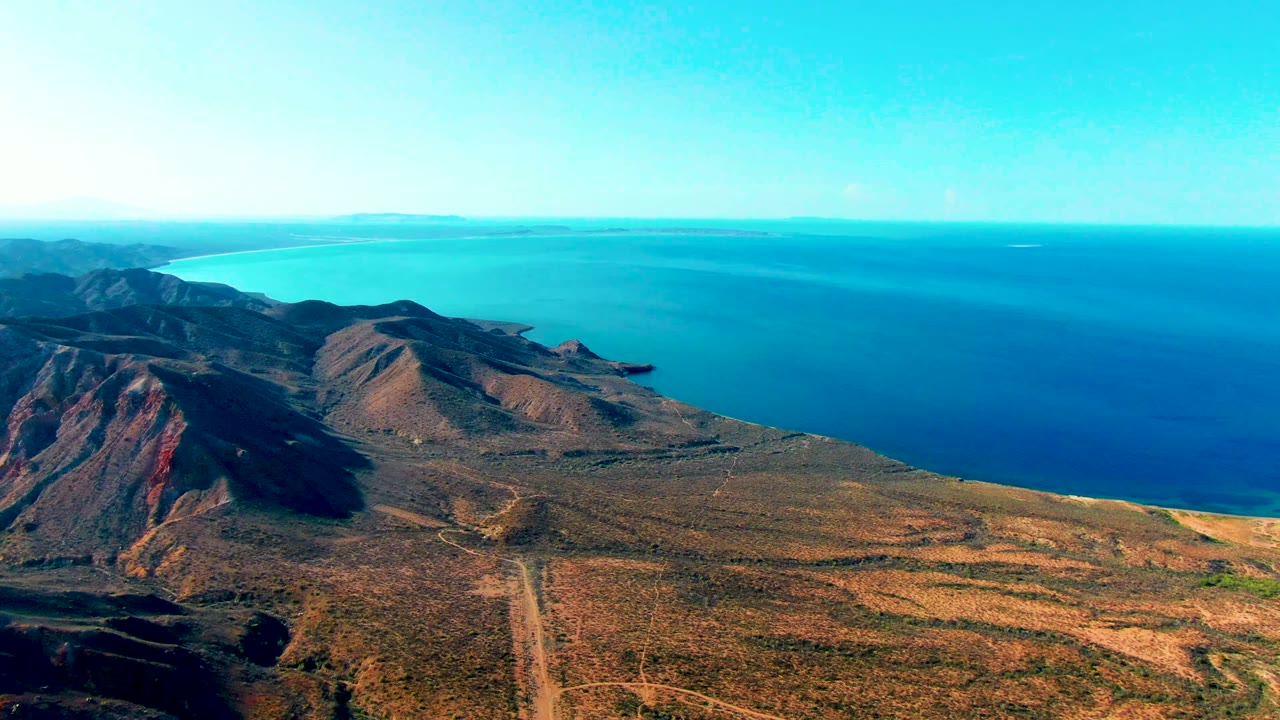 Opening aerial frame over Mulege, Baja California Sur, Mexico, showing dry hills, a winding dirt road, blue ocean, distant landforms, and no visible people.