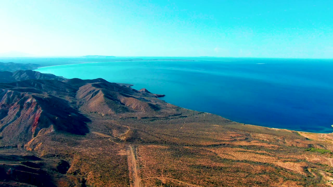 High aerial frame in Mulege, Baja California Sur, Mexico, with reddish-brown mountains, open arid ground, a narrow road, blue water beyond, and no visible people.