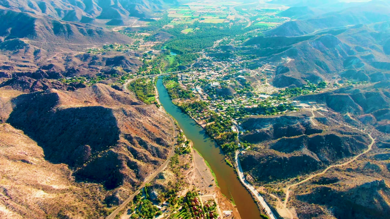 High aerial frame over Mulege, Baja California Sur, with a brown river curving past dry rocky slopes, green riverside vegetation, a small settlement, and no visible people.