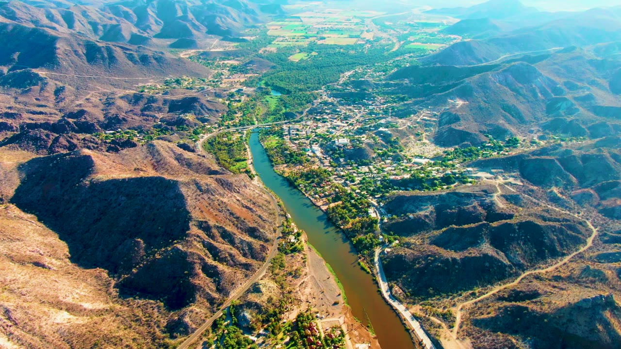High oblique frame over Mulege, Baja California Sur, showing the river cutting upward through the valley beside green banks, tan hills, roads, buildings, and no visible people.