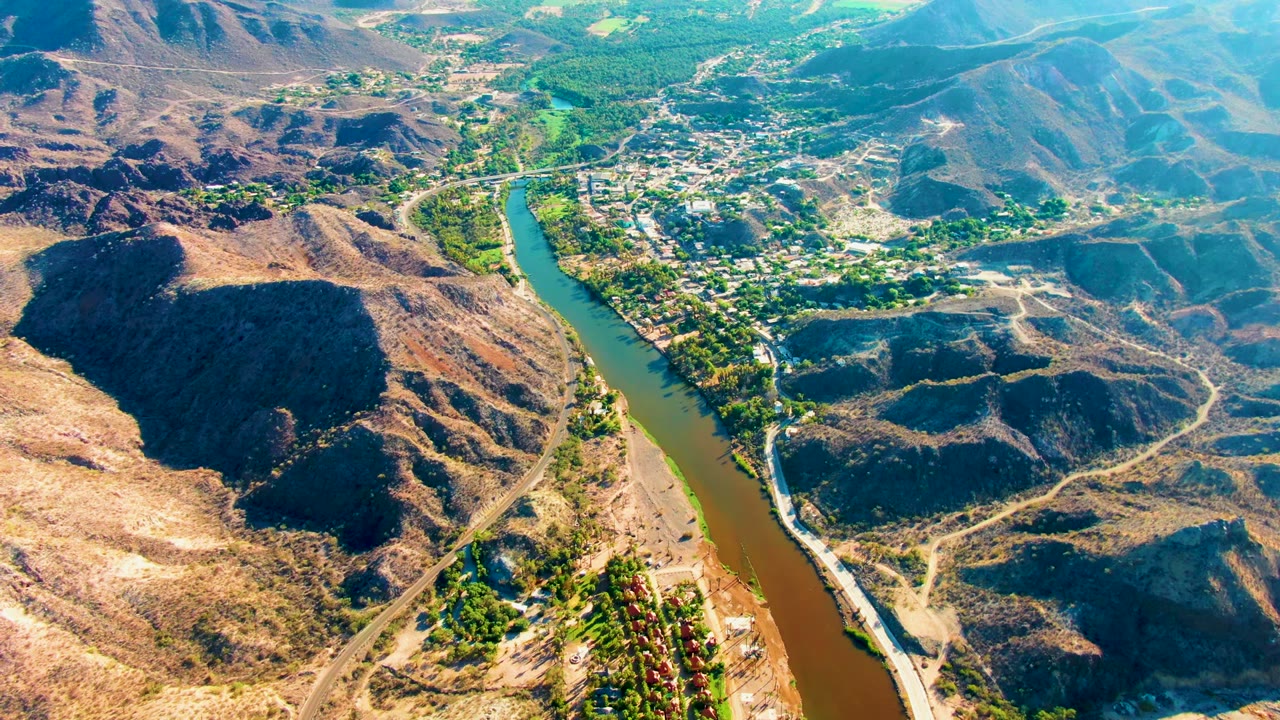 Aerial view over Mulege, Baja California Sur, with a green-brown river cutting between dry hills toward a small settlement, no visible people.