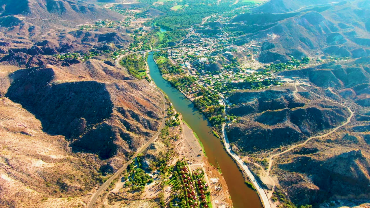 Drone-style view over Mulege, Baja California Sur, showing the river bending past dry ridges with clustered buildings and greenery in the upper valley, no visible people.