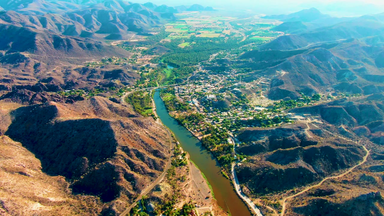 Opening aerial frame over Mulege, Baja California Sur, with a green-brown river curving past a small settlement, dry hills on both sides, and no visible people.