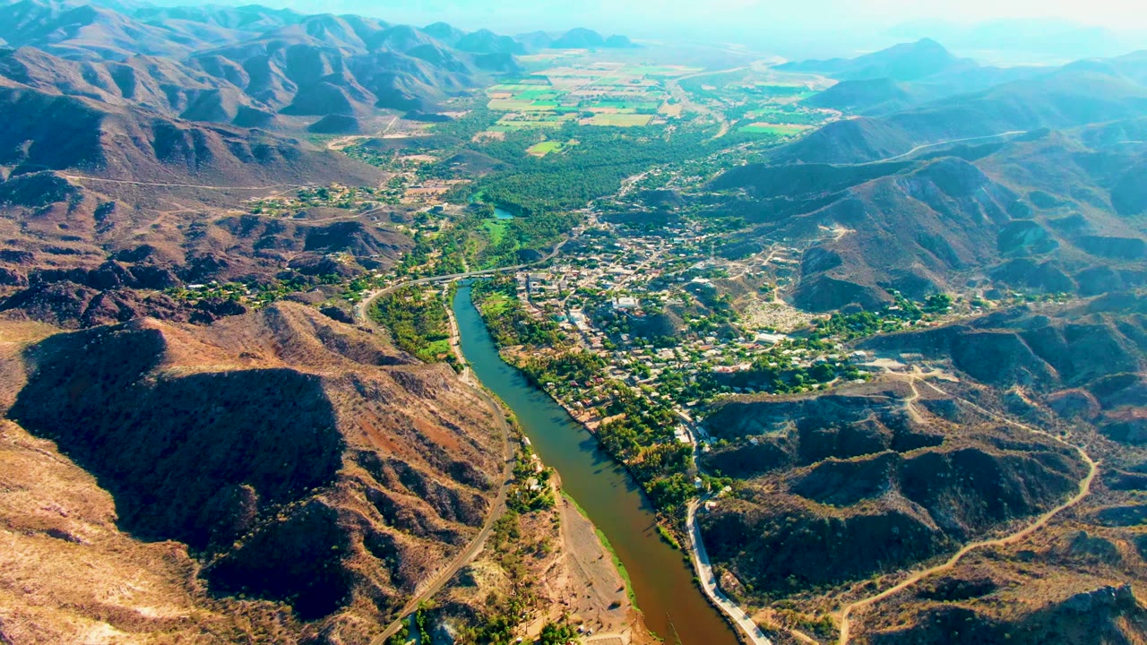 The aerial frame shows Mulege's river running through the valley, with the settlement and roads beside it, green patches beyond town, brown mountains, and no visible people.