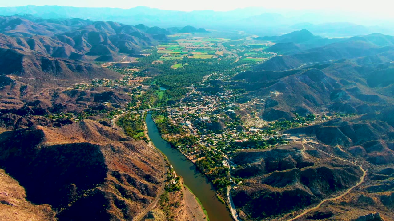 Aerial view over Mulege, Baja California Sur, with a green river bending past town buildings, dry mountain ridges, distant fields, hazy hills, and no visible people.