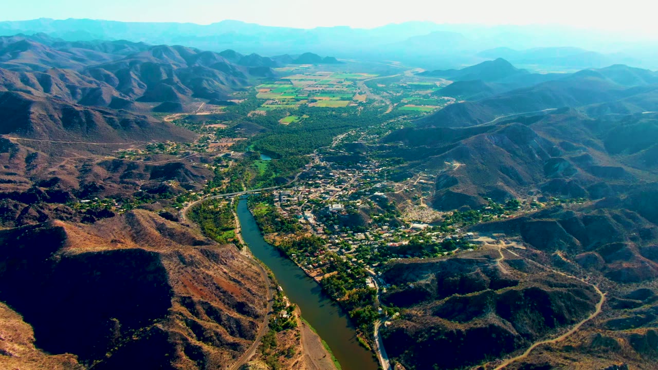 Aerial view across Mulege, Baja California Sur, with a river bend below, clustered settlement areas, dry ridges casting dark shadows, bright green fields in the distance, and no visible people.