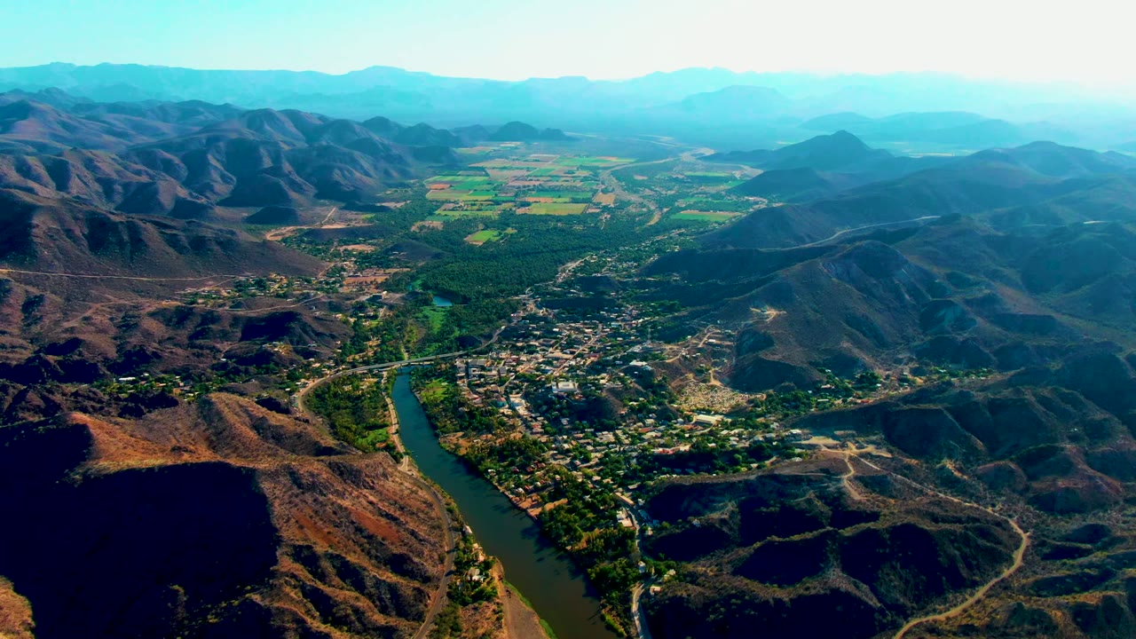 Aerial view over Mulege, Baja California Sur, Mexico, with a green river at lower left, a small town along the valley floor, brown ridges, green fields, hazy mountains, and no visible people.