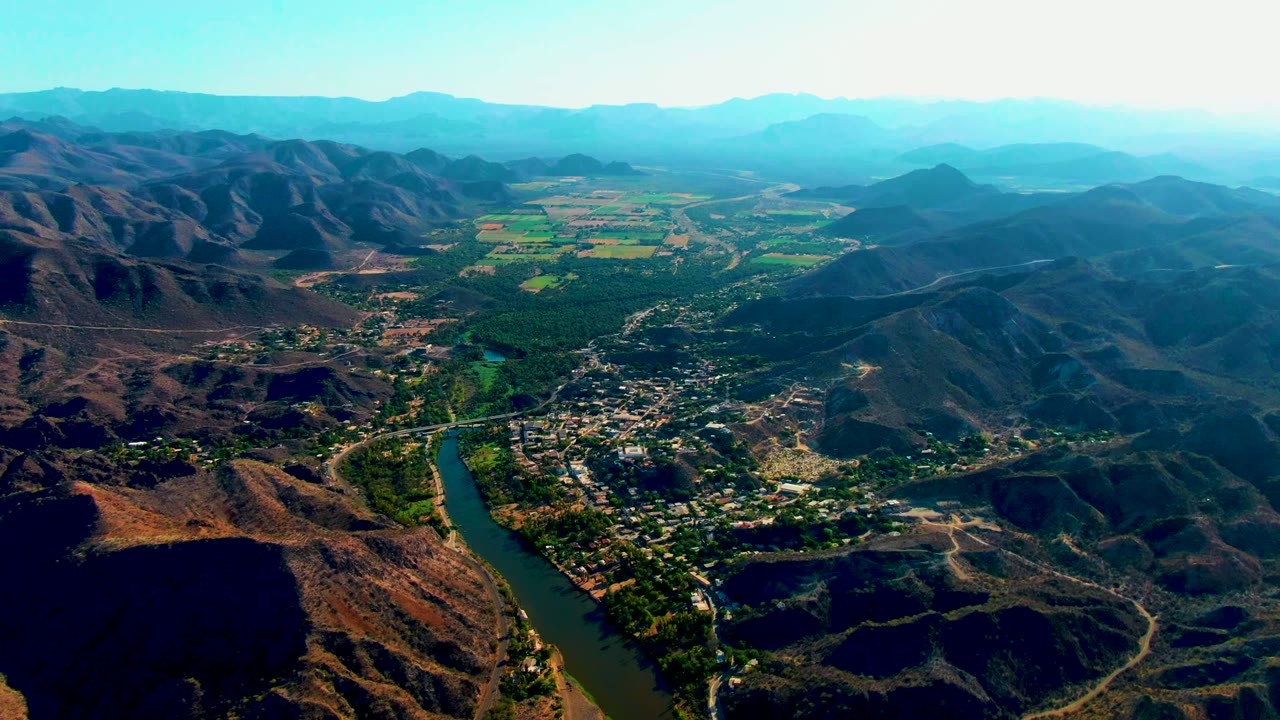 Aerial view of Mulege's river valley with the water channel at lower left, town blocks spreading across the lower center, dark ridge shadows, green fields beyond town, and no visible people.