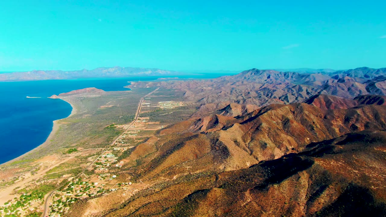 Aerial view over Mulege, Baja California Sur, with brown coastal mountains at right, blue ocean at left, sandy shoreline, scattered settlement, and no visible people.