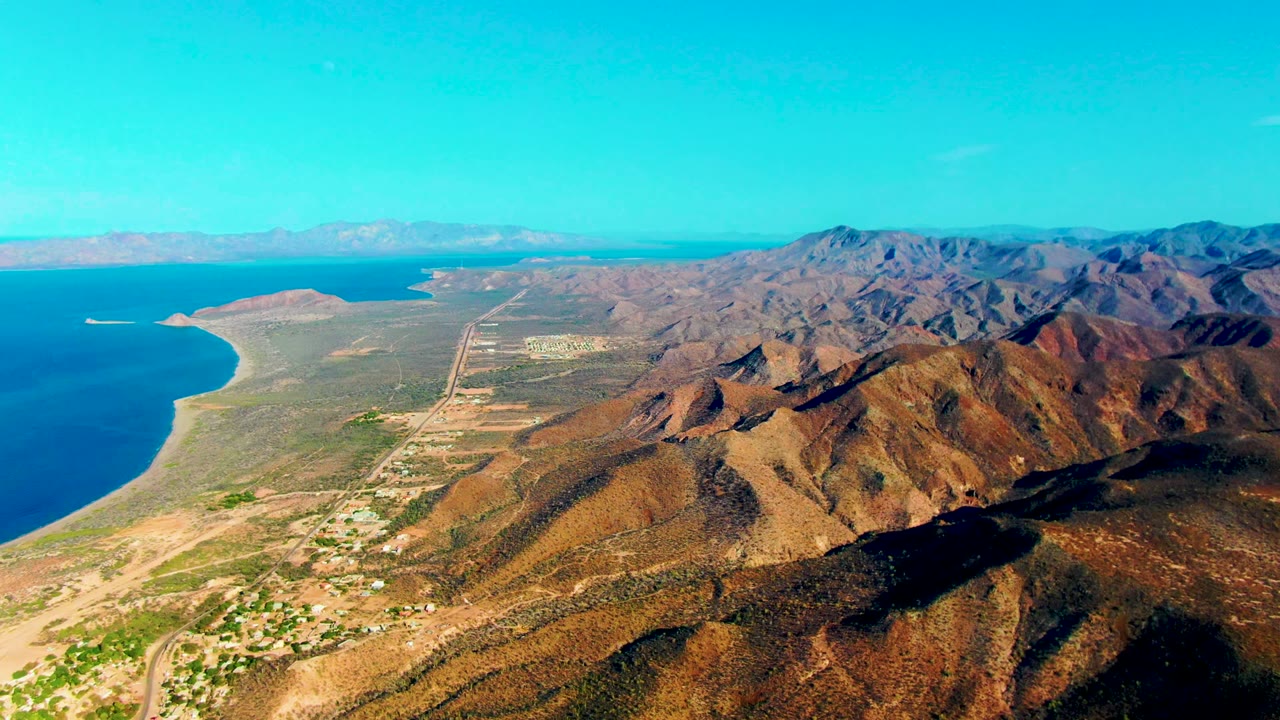 Wide aerial view of coastline, dry mountain range, roads, and small settlement.
