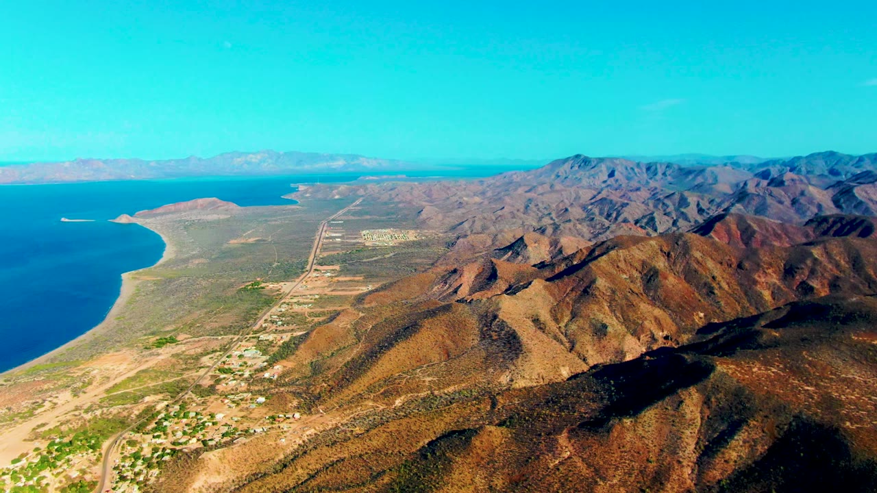Aerial view in Mulege with the blue water and sandy beach to the left, settlement roads through the center, sunlit brown hills to the right, and no visible people.