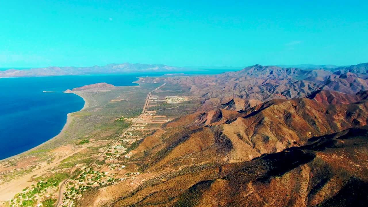 Aerial view over Mulege, Baja California Sur, with blue ocean on the left, a sandy curving shore, brown mountain ridges in the foreground, sparse development below, and no visible people.