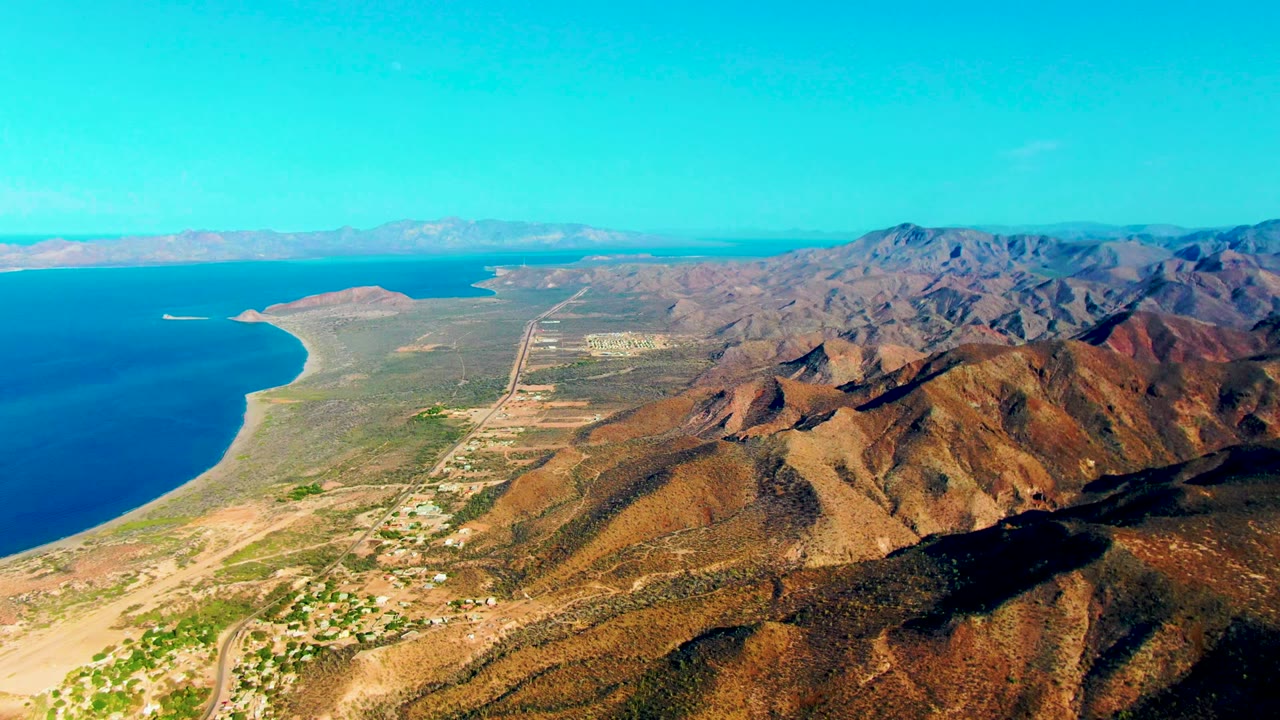 High coastal view with turquoise water, beach, desert hills, roads, and a small boat offshore.