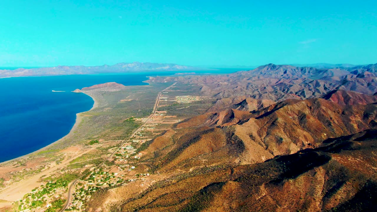Wide aerial view in Mulege, Baja California Sur, with open sea, a curved shoreline, an inland road grid, scattered structures, rugged brown mountains, and no visible people.