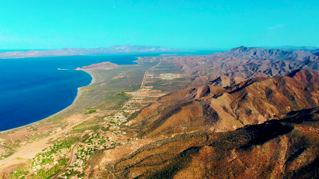 Opening aerial view over Mulege, Baja California Sur, with blue ocean at left, brown mountains at right, a runway corridor through the midground, and no visible people.