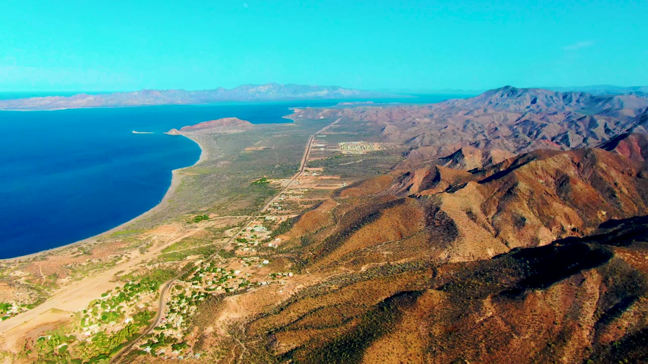 Mulege aerial view with the ocean bay on the left, a sandy point in the distance, runway and roads across the center, mountains on the right, and no visible people.