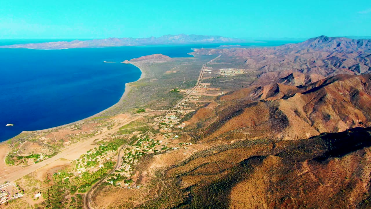 Opening aerial frame over Mulege, Baja California Sur, showing blue ocean along a curved beach, arid hills, roads, scattered buildings, distant shoreline, and no visible people.