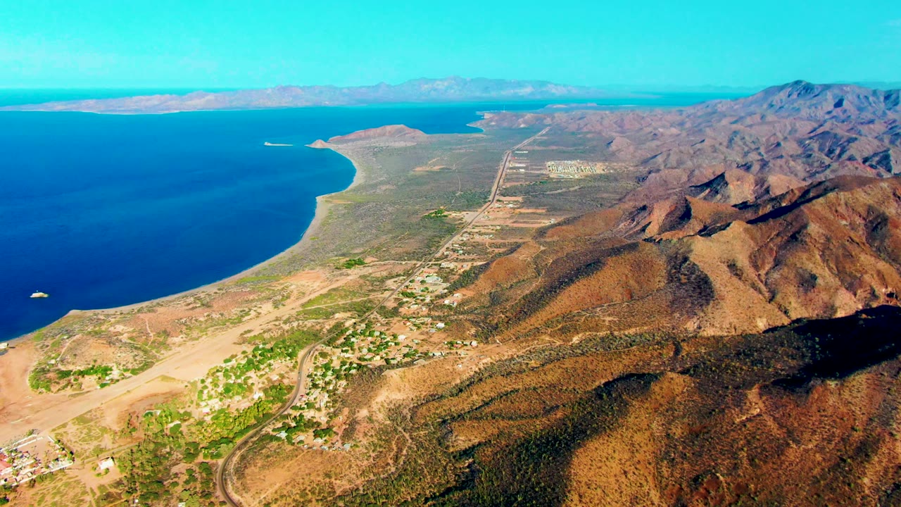 Second frame over Mulege coastline with the ocean occupying the left side, a pale beach below, road lines and settlement areas in the middle distance, mountains to the right, and no visible people.