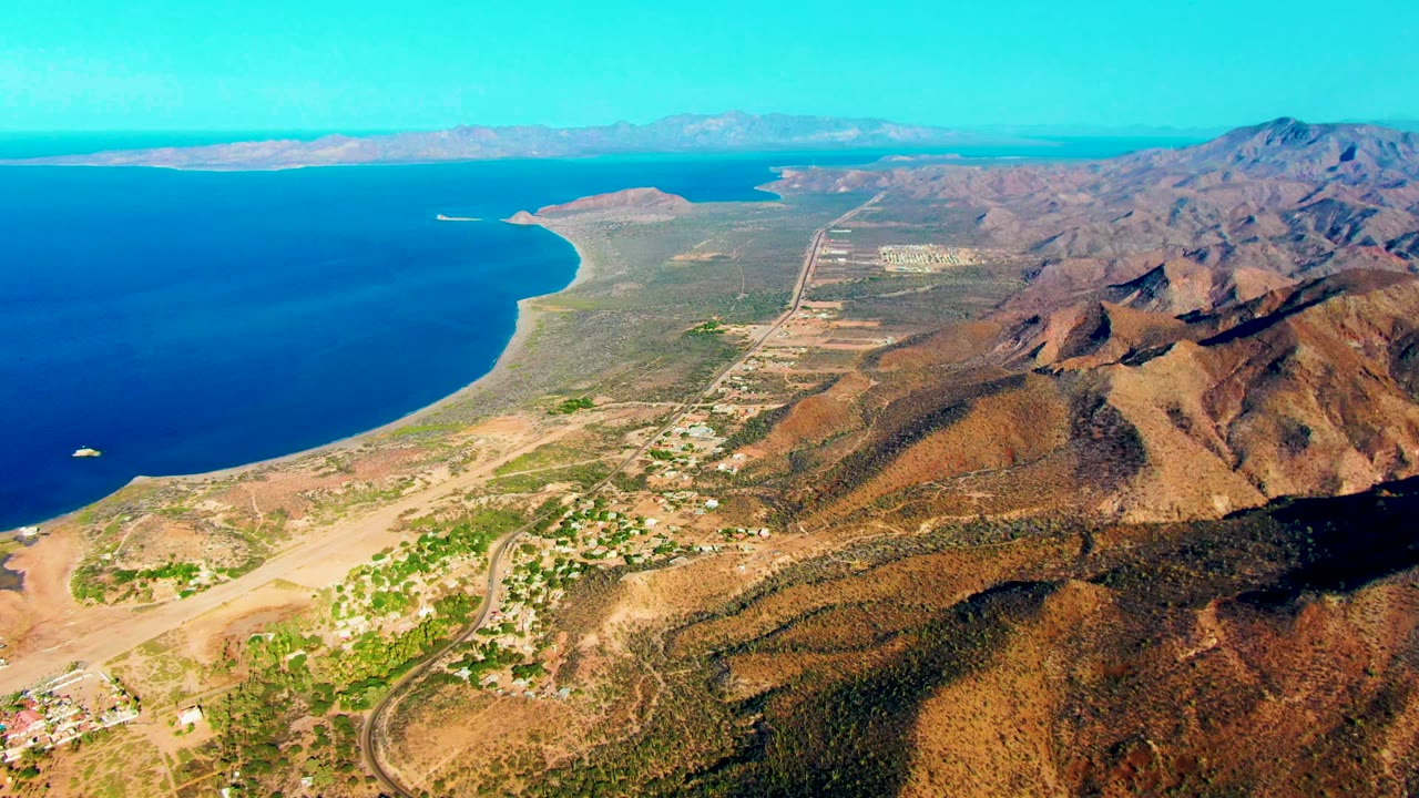 Third frame of the Mulege aerial shows a broad sweep of dark blue water, shoreline, dry brown ridges, pockets of greenery, small settlements near the lower edge, and no visible people.