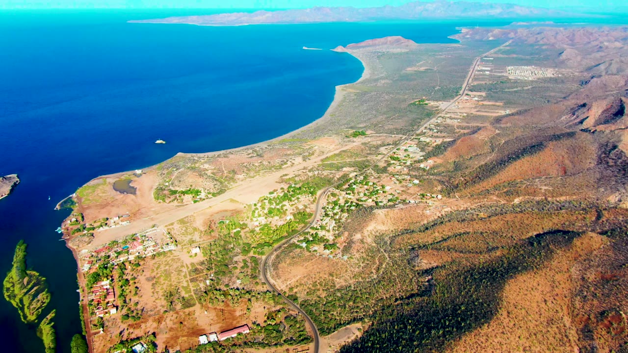 Opening aerial frame over Mulege, Baja California Sur, with deep blue water, dry hillside terrain, scattered buildings, a curving road, and no visible people.