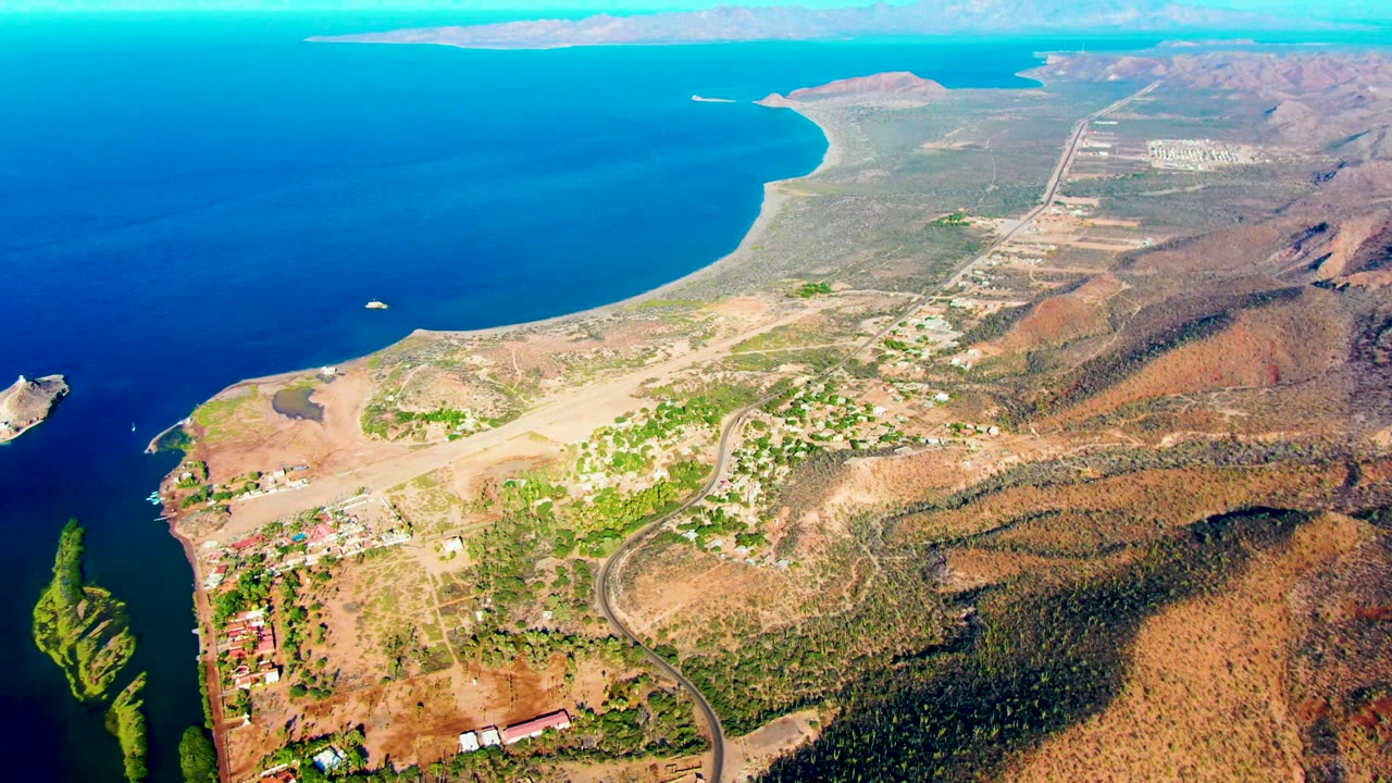 High aerial coastal view with ocean, hillside terrain, roads, and scattered buildings.