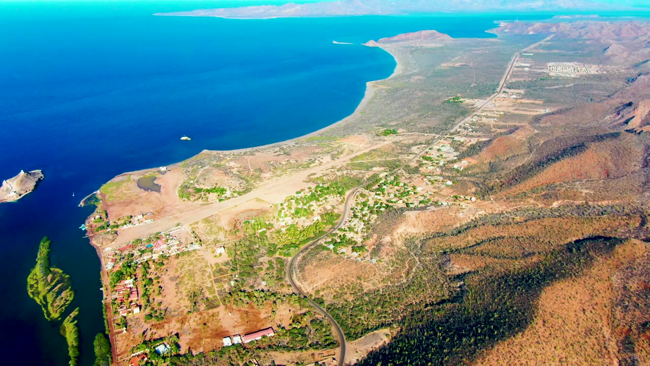 Drone-height frame over Mulege, Baja California Sur, showing a broad bay, reddish-brown hills, green vegetation pockets, shoreline buildings, and no visible people.