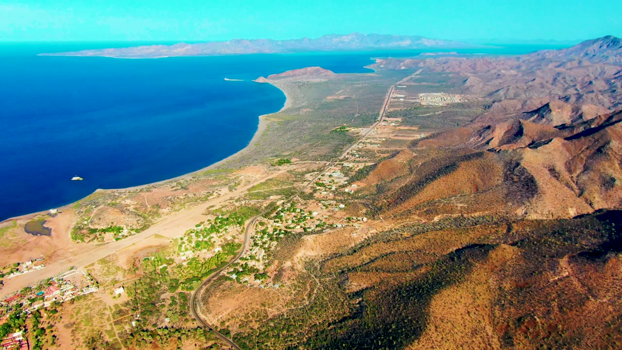 High aerial frame over Mulege, Baja California Sur, showing deep blue ocean, a curving tan shoreline, dry hills, scattered buildings, roads, distant land, and no visible people.
