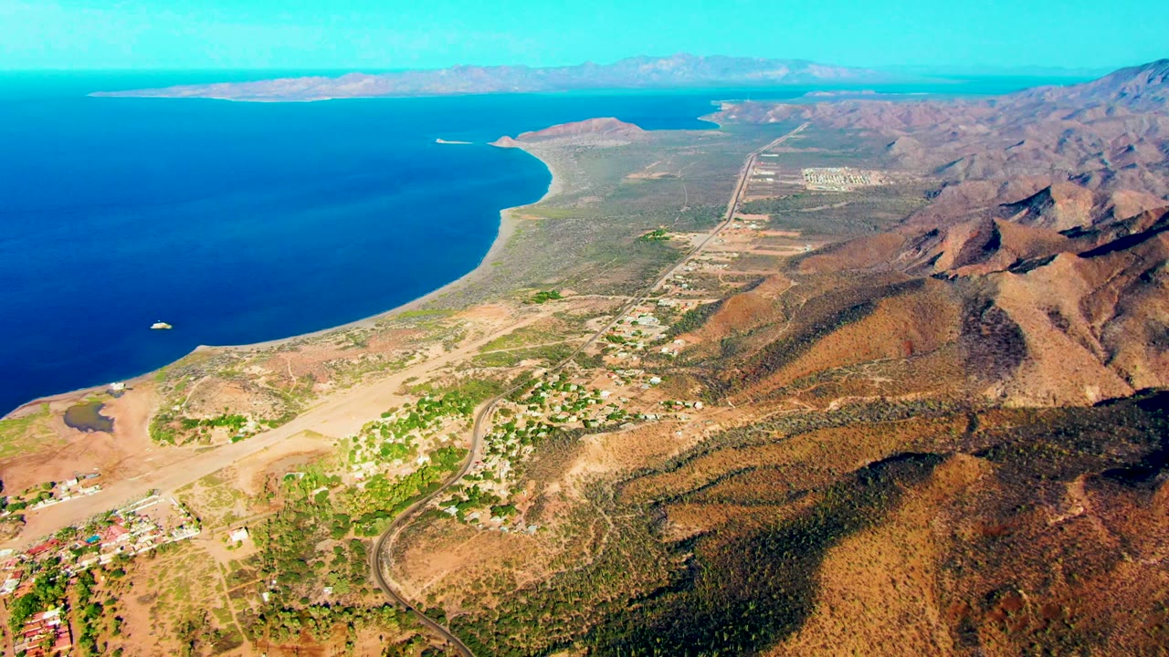 Aerial coastline with a small boat offshore, settlement roads, beach, and mountains.
