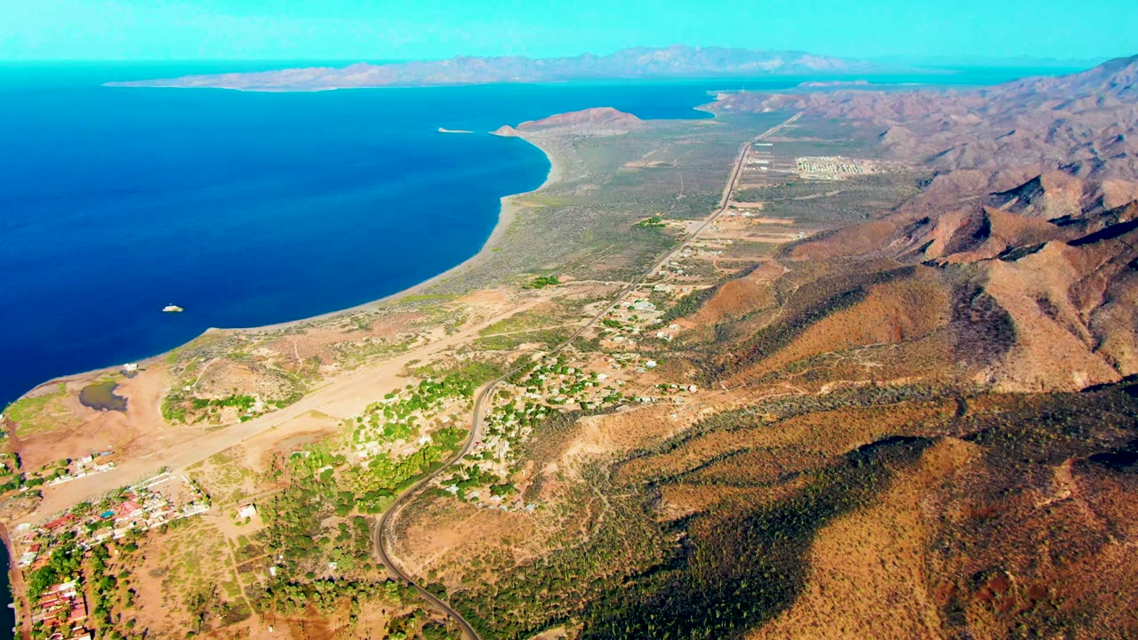 Aerial frame over Mulege with ocean filling the left side, a rounded coastal point and beach below, inland roads and small buildings in the middle distance, brown mountain slopes to the right, and no visible people.