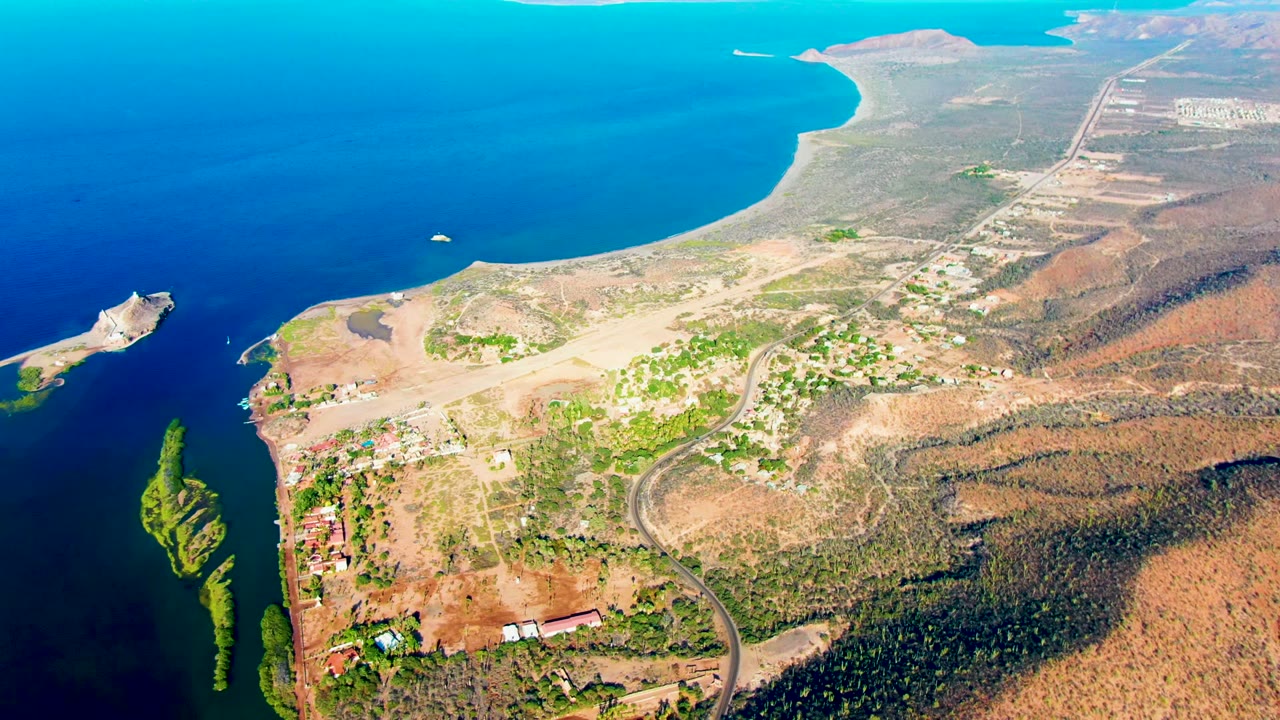 Early aerial frame over Mulege, Baja California Sur shows deep blue ocean, a greenish inlet, a small pier or breakwater, roads, scattered buildings, dry hills, and no visible people.