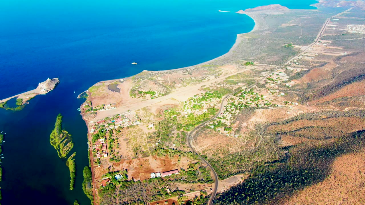 At 0:02, the Mulege aerial view holds the curved blue bay at the top, the greenish inlet and small pier to the left, inland roads and buildings below, and no visible people.