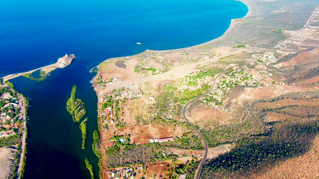 Aerial frame over Mulege, Baja California Sur showing deep blue water at left and top, dry shoreline land, green patches, a winding road, a small white boat near open water, and no visible people.