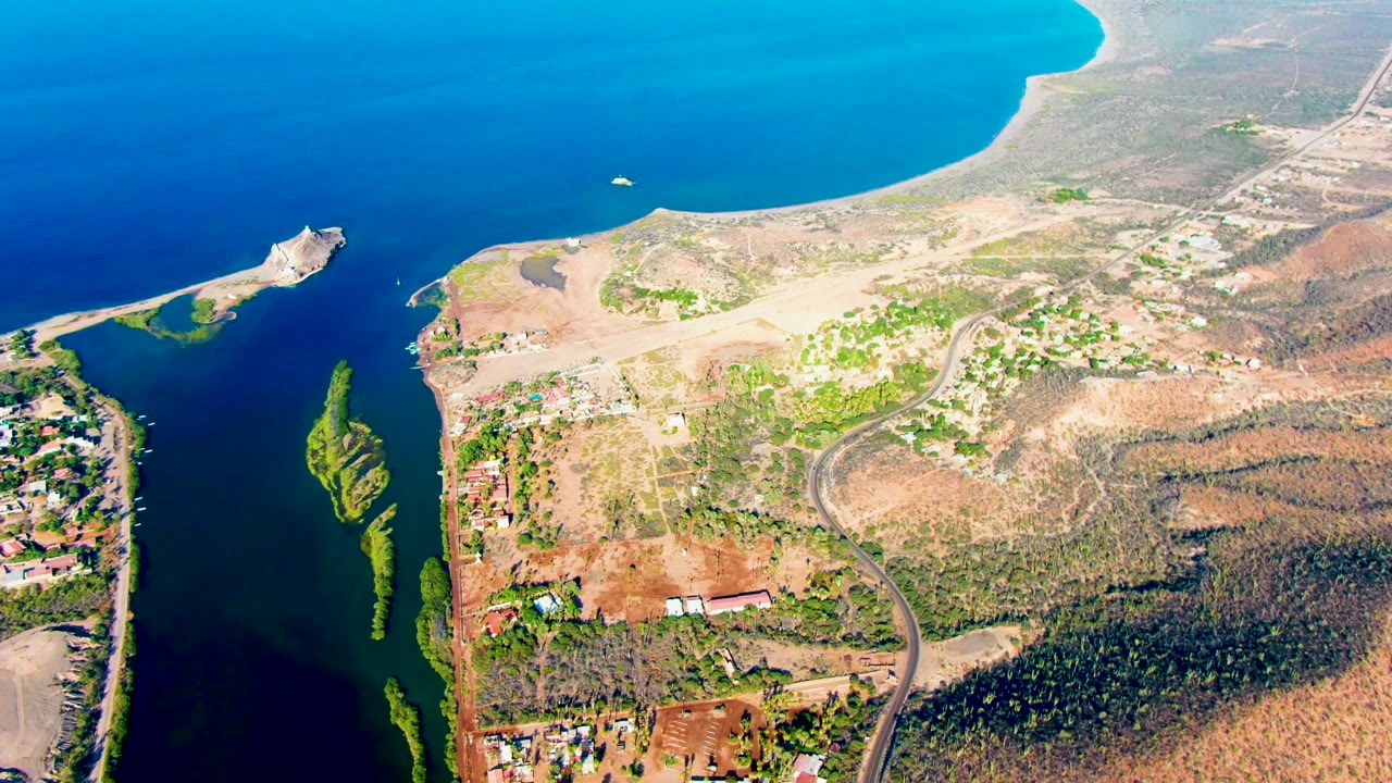 Drone-style aerial view of a coastal waterway with a small boat, arid land, road, and green patches.