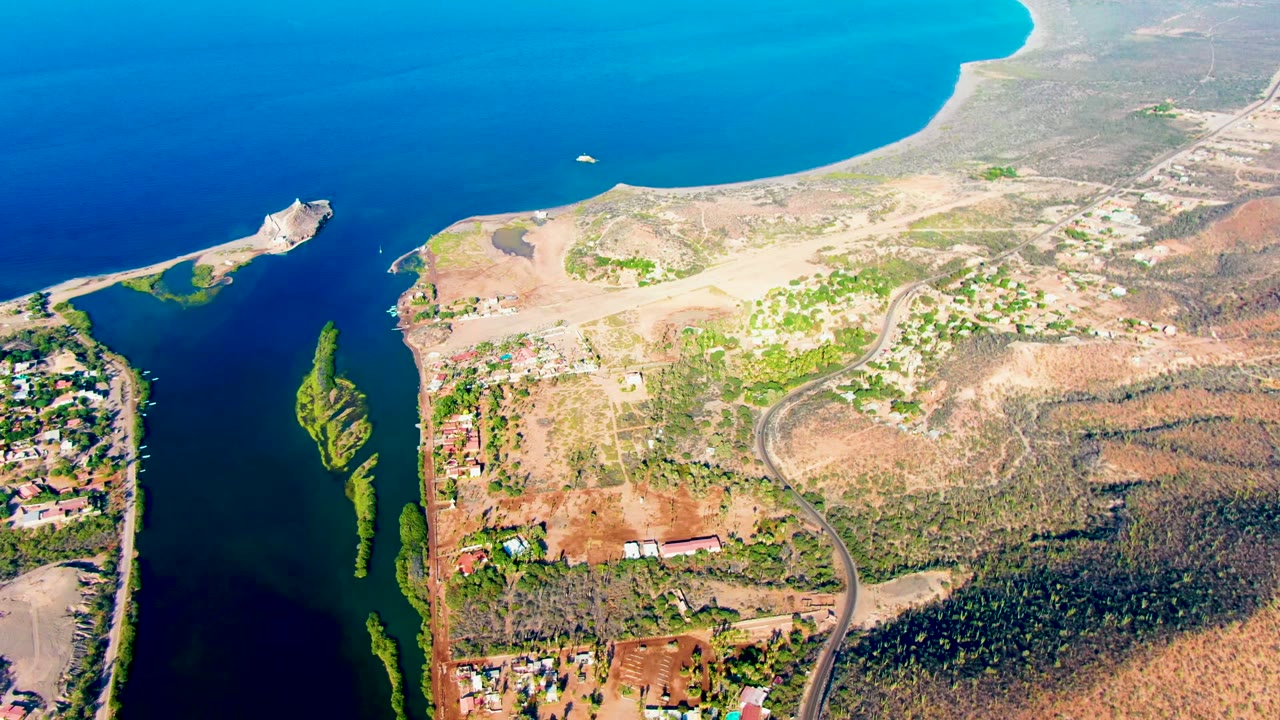 Aerial frame over Mulege, Baja California Sur showing the boat continuing across the upper water area, with narrow green-edged channels below, dry land to the right, and no visible people.