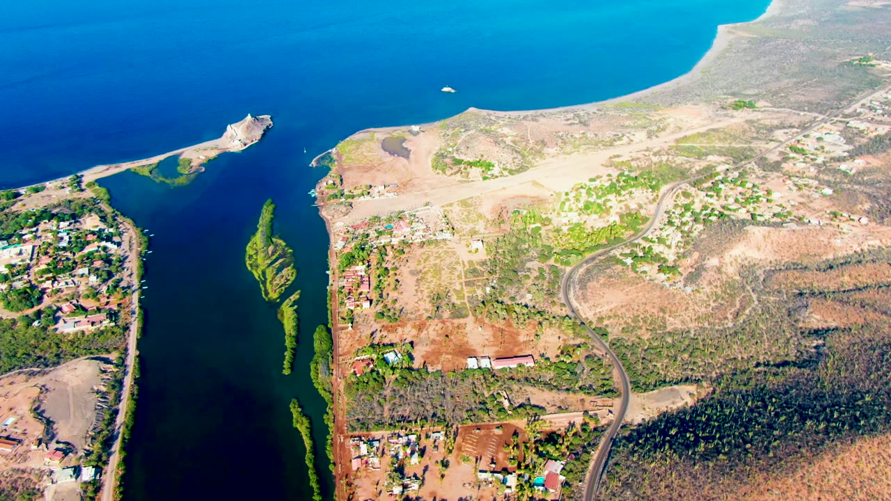 High aerial view over Mulege, Baja California Sur, showing a blue inlet, narrow green vegetation strips, dry development land, scattered buildings, and no visible people.