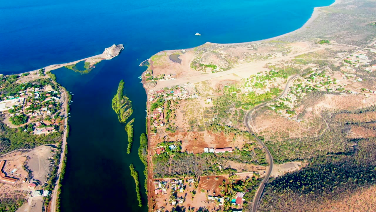 High drone perspective of Mulege, Baja California Sur, with the inlet cutting along the left side, exposed construction land and scrub on the right, buildings near the lower edge, and no visible people.