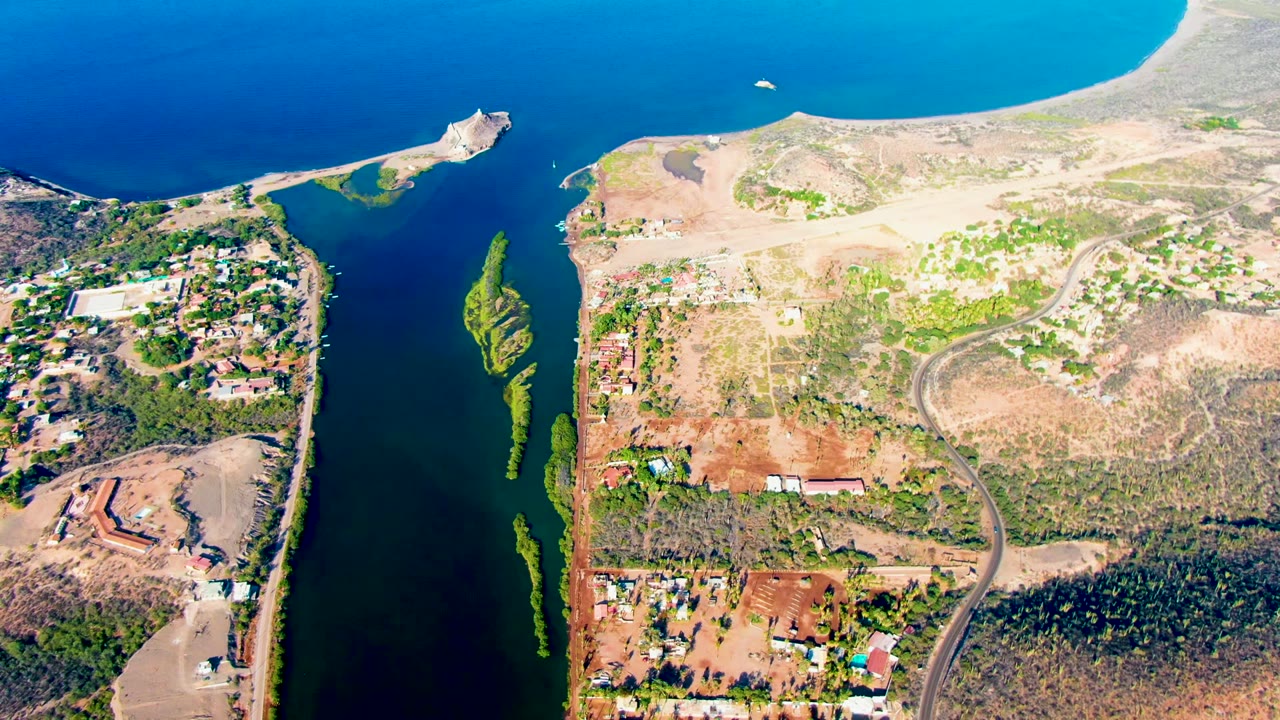 Aerial frame over Mulege, Baja California Sur, showing a dark water channel between landmasses, a breakwater area, dry terrain, roads, scattered buildings, bright blue ocean, a small boat near the upper water, and no visible people.
