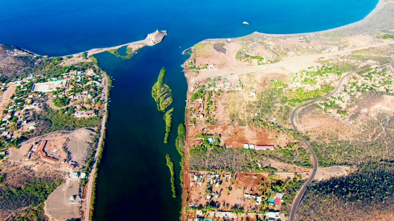 Aerial frame over Mulege, Baja California Sur, showing the narrow channel and blue ocean with a pale sand edge, low buildings, a curving road, green water vegetation, and no visible people.
