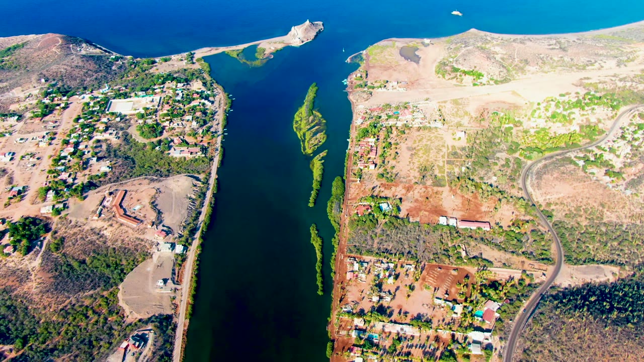 High aerial frame of Mulege showing a vertical strip of green algae in the water, a curved road through dry terrain, scattered structures, hills, and no visible people.