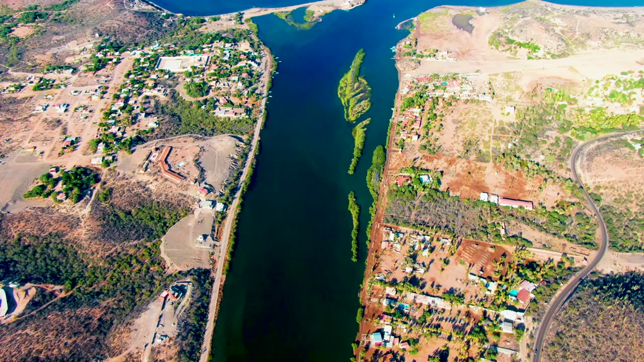 High aerial frame over Mulege, Baja California Sur, with blue-green river water beside dry shoreline, scattered buildings, roads, and no visible people.