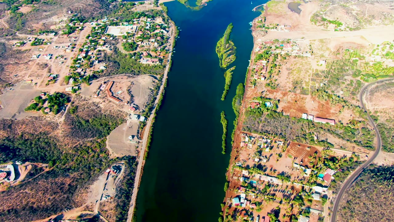 Overhead frame from Mulege with dark river water, a narrow green vegetated strip in the channel, dry brown hillsides, scattered buildings, and no visible people.