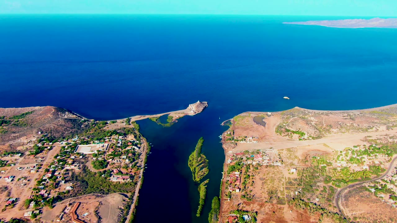 Aerial frame over Mulege, Baja California Sur, with a dark canal beside a small settlement, open dry land to the right, green algae in the water, blue sea beyond, and no visible people.
