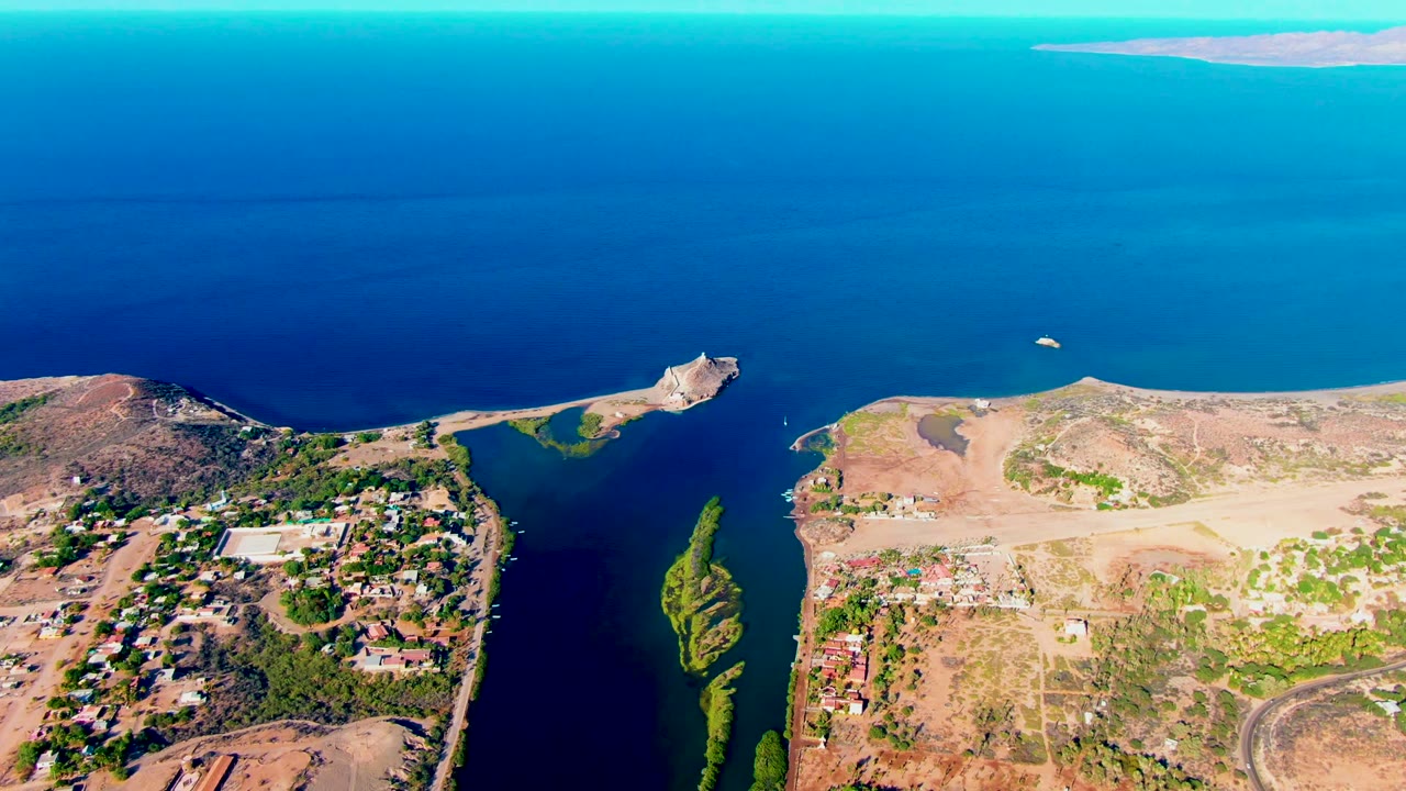 Aerial frame in Mulege with the canal mouth and algae patches visible below, a small settlement at left, dry peninsula terrain at right, bright blue water beyond, and no visible people.