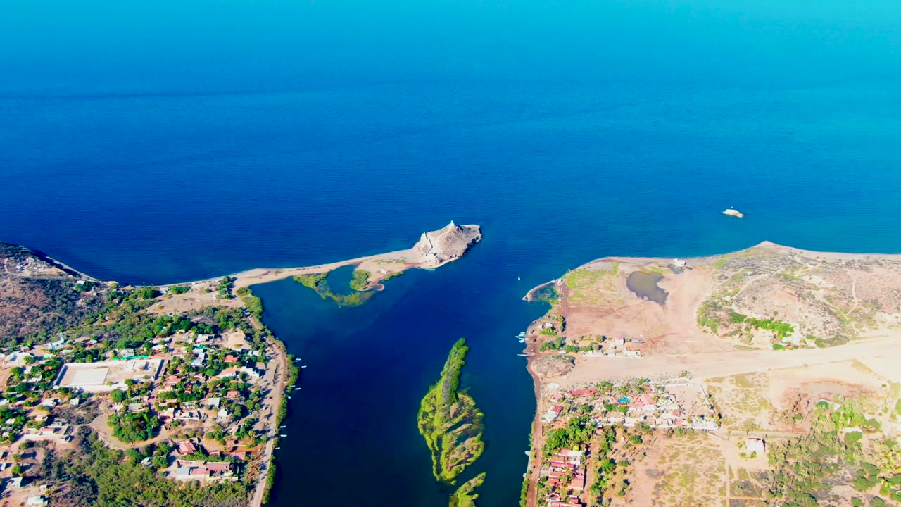 Aerial view over Mulege, Baja California Sur, with blue water, a pale shoreline, dry terrain, scattered buildings, and no visible people.