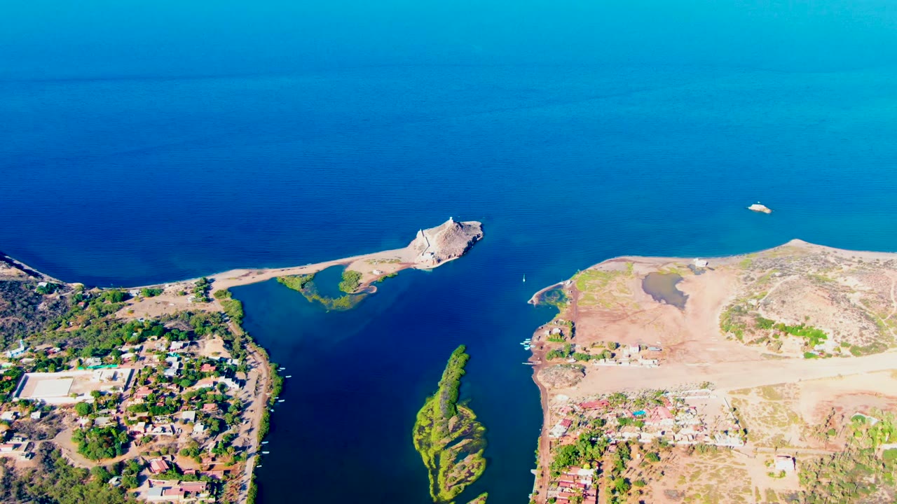 Aerial view in Mulege, Baja California Sur, with a small green island or marsh strip in dark blue water, dry coastal ground, buildings, and no visible people.