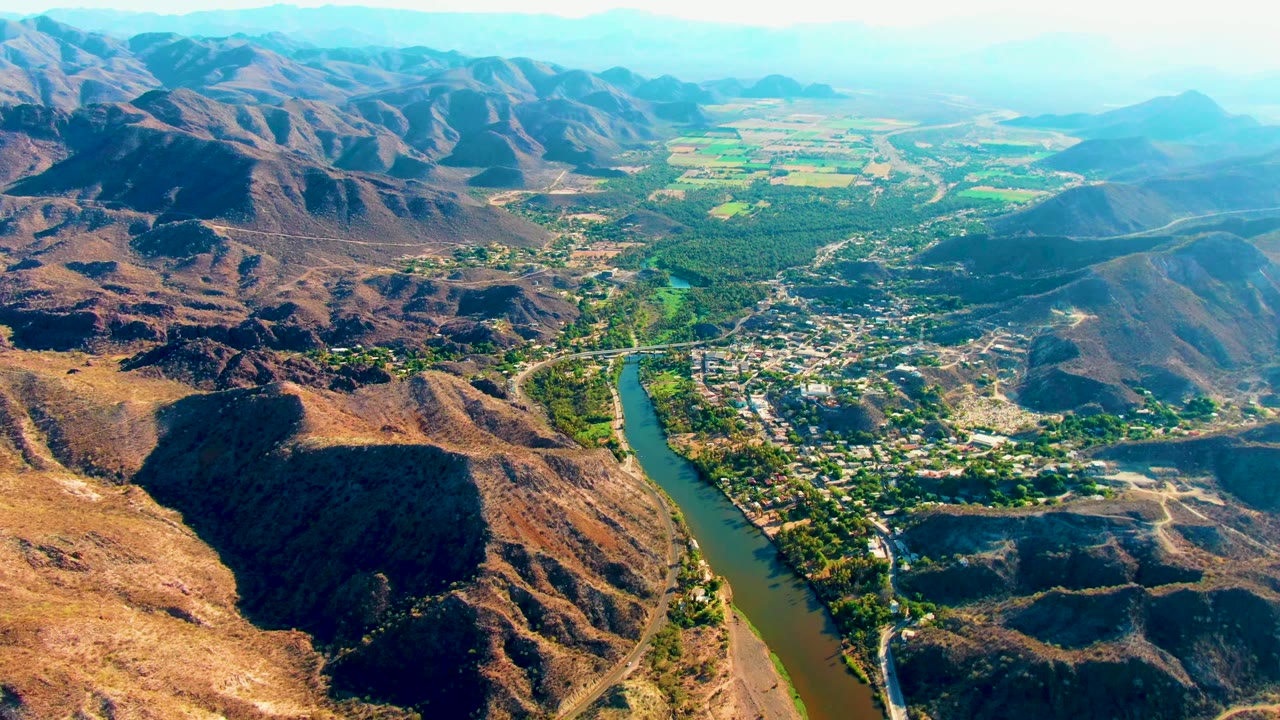 Aerial view over Mulege, Baja California Sur, showing a green-brown river curving past a small town, dry hills in the foreground, distant fields, hazy mountains, and no visible people.