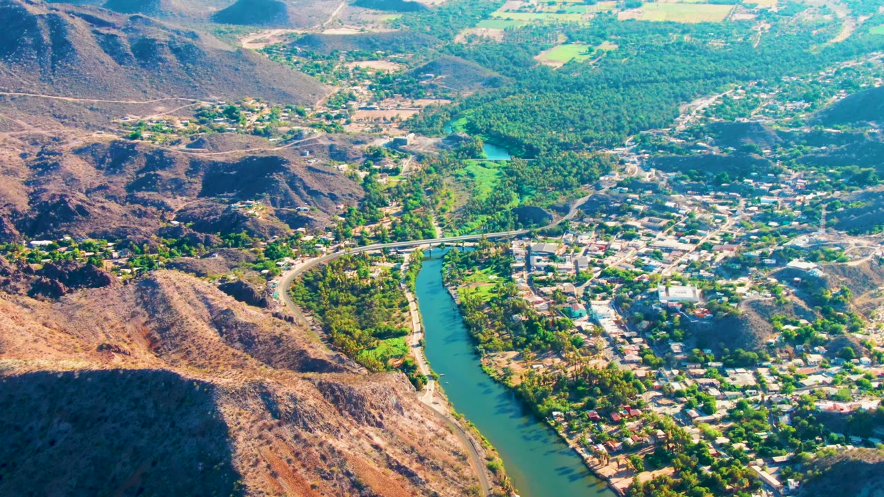 Aerial view over Mulege, Baja California Sur with a green river curving beside town buildings, palms, rocky hills, and no visible people.